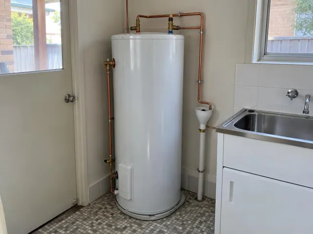 Cylindrical hot water tank beside a laundry sink and tiled wall in a small home interior in Mascot